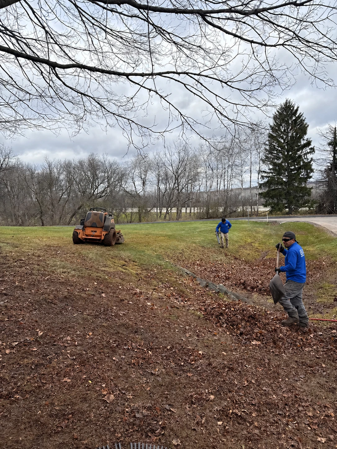 Two workers gathering autumn leaves on a sloped lawn beside a mower